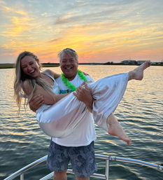 Laughing couple on a boat at sunset — man in shorts and a green lei lifting a woman in a shiny silver dress, barefoot, over calm coastal bay waters with a colorful sky.