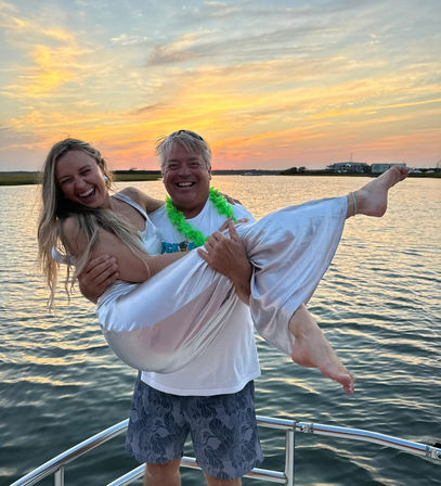 Laughing couple on a boat at sunset — man in shorts and a green lei lifting a woman in a shiny silver dress, barefoot, over calm coastal bay waters with a colorful sky.
