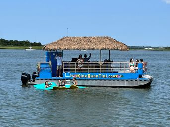 Blue tiki-style pontoon boat with a thatched roof, people waving and sunbathing, and friends lounging on a bright paddle float nearby in a sunny coastal bay.