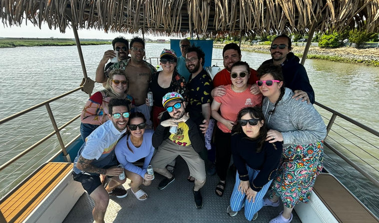 Group of friends smiling and holding drinks under a thatched-roof on a pontoon boat cruising a sunny river or coastal bay with marshy shoreline