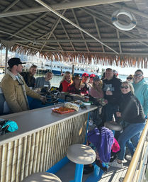 Smiling group enjoying drinks and snacks at a bamboo tiki-bar on a pontoon boat in a sunny marina, with a ferry and docks visible in the harbor.