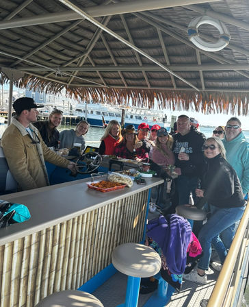 Smiling group enjoying drinks and snacks at a bamboo tiki-bar on a pontoon boat in a sunny marina, with a ferry and docks visible in the harbor.