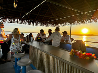 Tiki-style boat bar with bamboo counter and thatched roof, passengers wearing colorful leis enjoy a coastal sunset cruise over calm bay waters with the sun setting on the horizon.