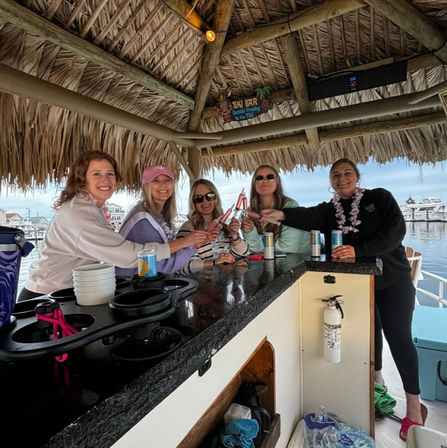Five friends smiling and clinking canned drinks at a thatched tiki-roof bar aboard a boat docked at a marina, with yachts and a cloudy sky in the background.