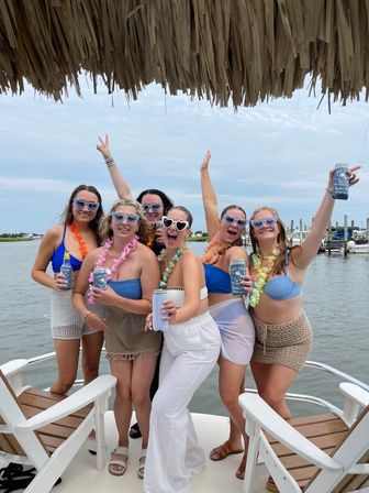 Six friends in heart-shaped sunglasses and leis enjoying a summer boat party, holding canned drinks under a tiki thatch with marina docks in the background.