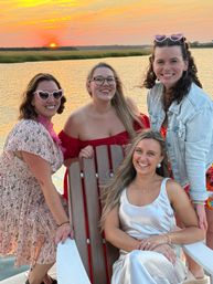 Four friends on a dock at a marshy waterfront during a golden sunset, smiling in summer dresses and heart-shaped sunglasses, one seated in an Adirondack chair.