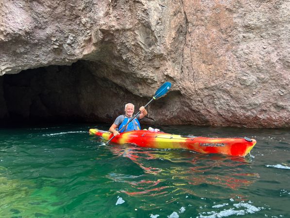 Kayaker paddling a red-and-yellow kayak into a rocky coastal sea cave with rugged pinkish cliffs and clear emerald-green water, outdoor kayaking adventure.