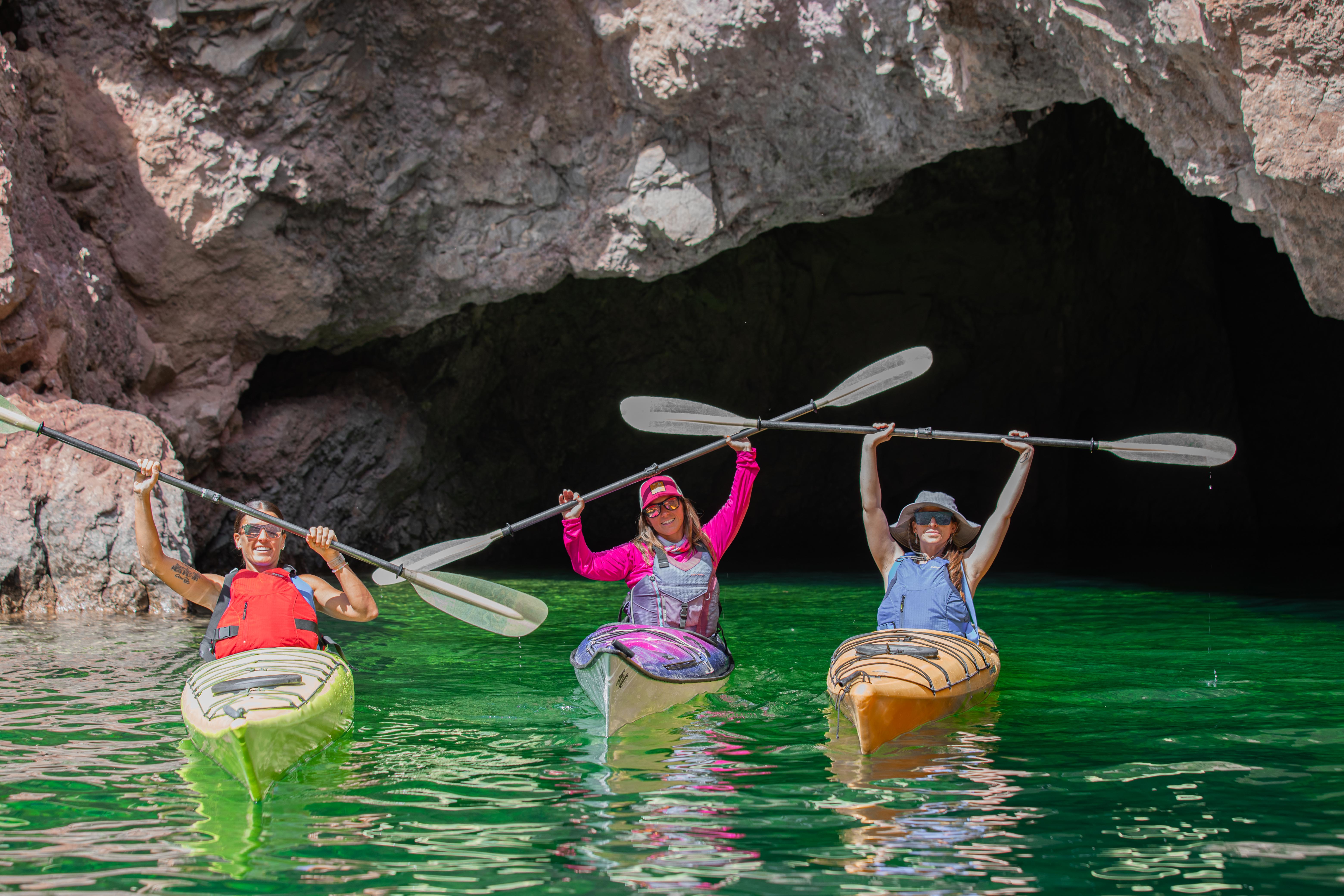 Three kayakers in colorful kayaks holding paddles overhead in front of a rocky sea cave on vivid emerald-green water — coastal kayaking adventure.
