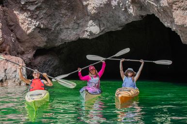 Three kayakers in colorful kayaks holding paddles overhead in front of a rocky sea cave on vivid emerald-green water — coastal kayaking adventure.