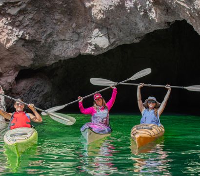 Three kayakers in colorful kayaks holding paddles overhead in front of a rocky sea cave on vivid emerald-green water — coastal kayaking adventure.