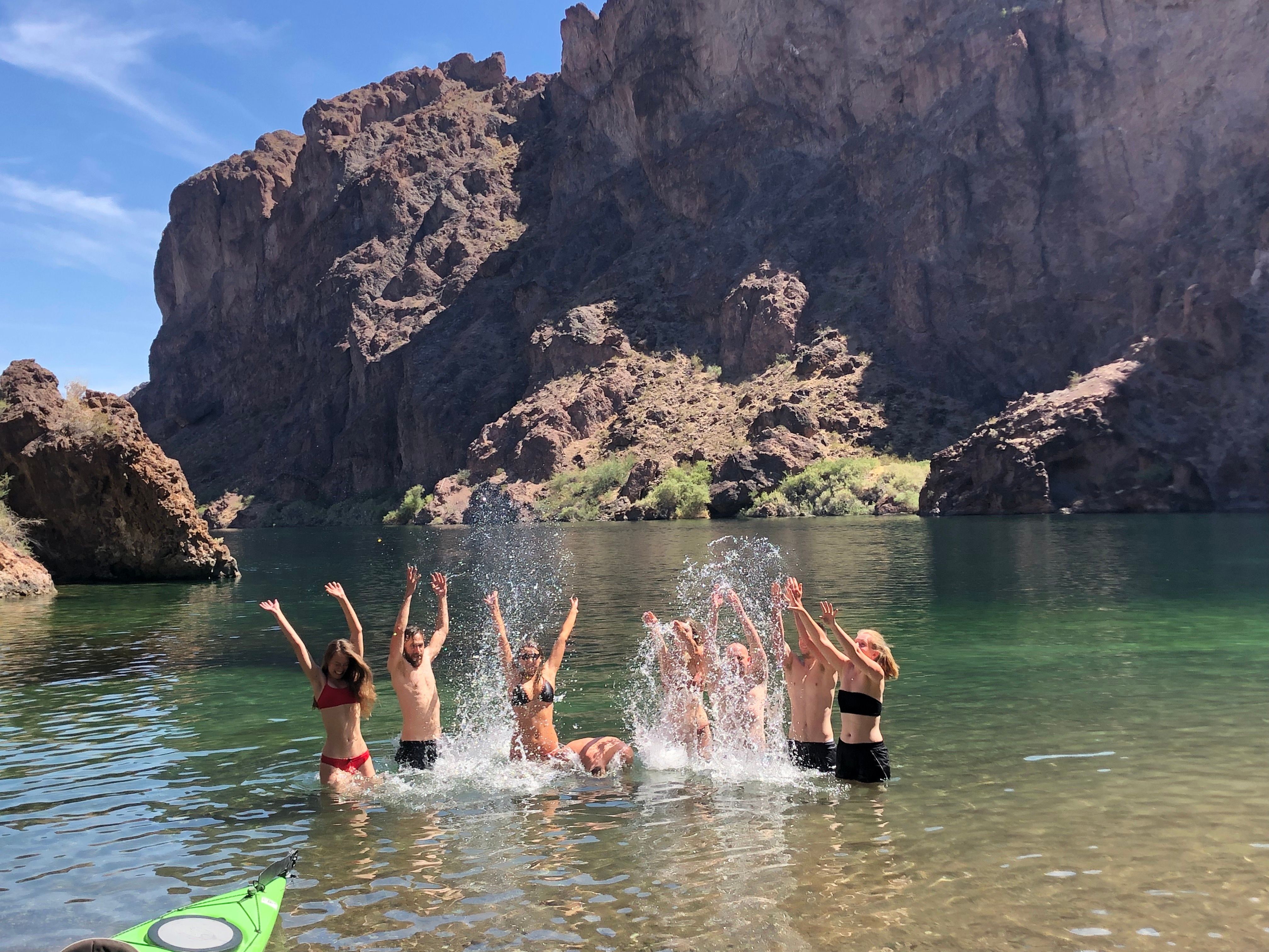 Group of people splashing and cheering in a clear green canyon lake with towering rocky cliffs in the background and a green kayak in the shallow foreground.