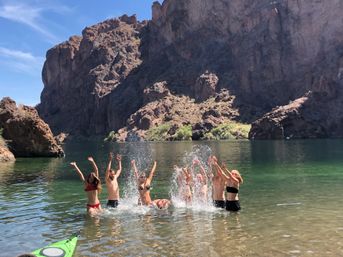 Group of people splashing and cheering in a clear green canyon lake with towering rocky cliffs in the background and a green kayak in the shallow foreground.