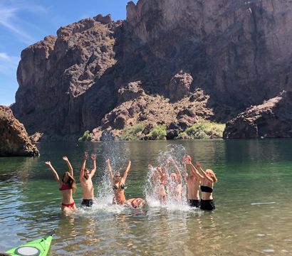 Group of people splashing and cheering in a clear green canyon lake with towering rocky cliffs in the background and a green kayak in the shallow foreground.