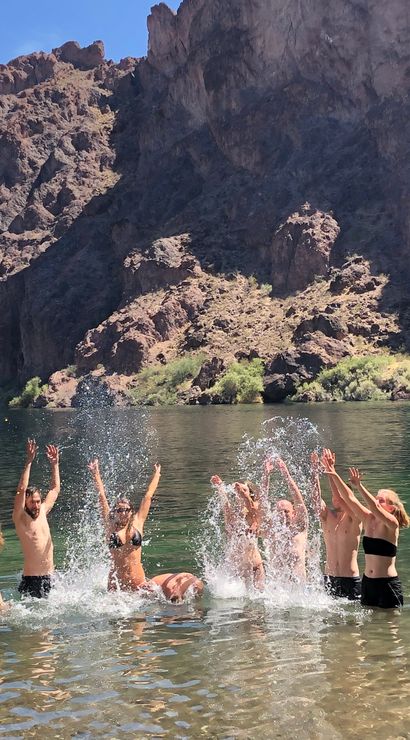 Group of people splashing and cheering in a clear green canyon lake with towering rocky cliffs in the background and a green kayak in the shallow foreground.