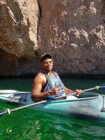 Smiling paddler in a turquoise kayak wearing sunglasses and a life jacket flashes a peace sign beside a sunlit rocky canyon wall over emerald-green water.