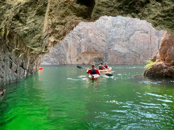 Kayakers paddling through a rocky cave arch into a sunlit canyon over emerald-green water