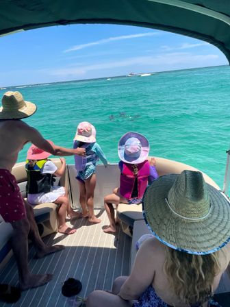 Family on a shaded pontoon boat watching dolphins in clear turquoise coastal waters — kids in sun hats and life jackets, adults wearing straw hats under a blue sky.