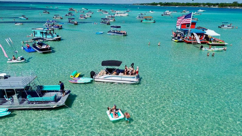 Aerial view of a sunny sandbar party with dozens of pontoon and party boats anchored in crystal-clear turquoise shallow water, people on floats and paddleboards, and a large boat flying an American flag.