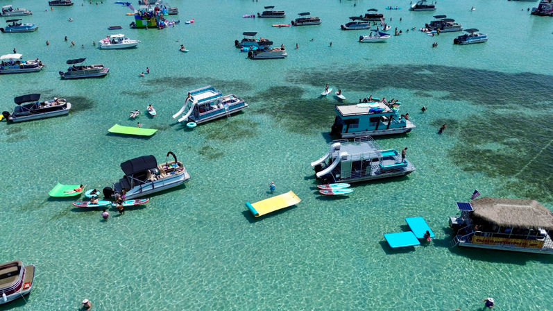 Aerial view of a busy sandbar in clear turquoise shallow water with pontoon boats, floating platforms and inflatables, people swimming, paddleboarding and lounging.