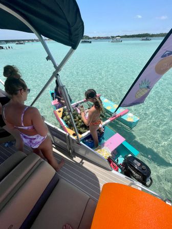 Women in swimsuits on a pontoon boat next to a colorful floating pineapple stand in crystal-clear turquoise tropical lagoon with anchored boats on the horizon
