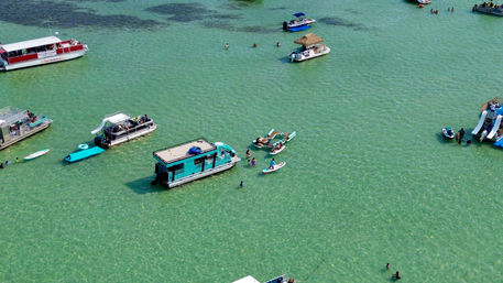 Aerial view of a sunny shallow turquoise lagoon dotted with houseboats, pontoons, paddleboards and small boats, with people swimming and relaxing on crystal-clear water.