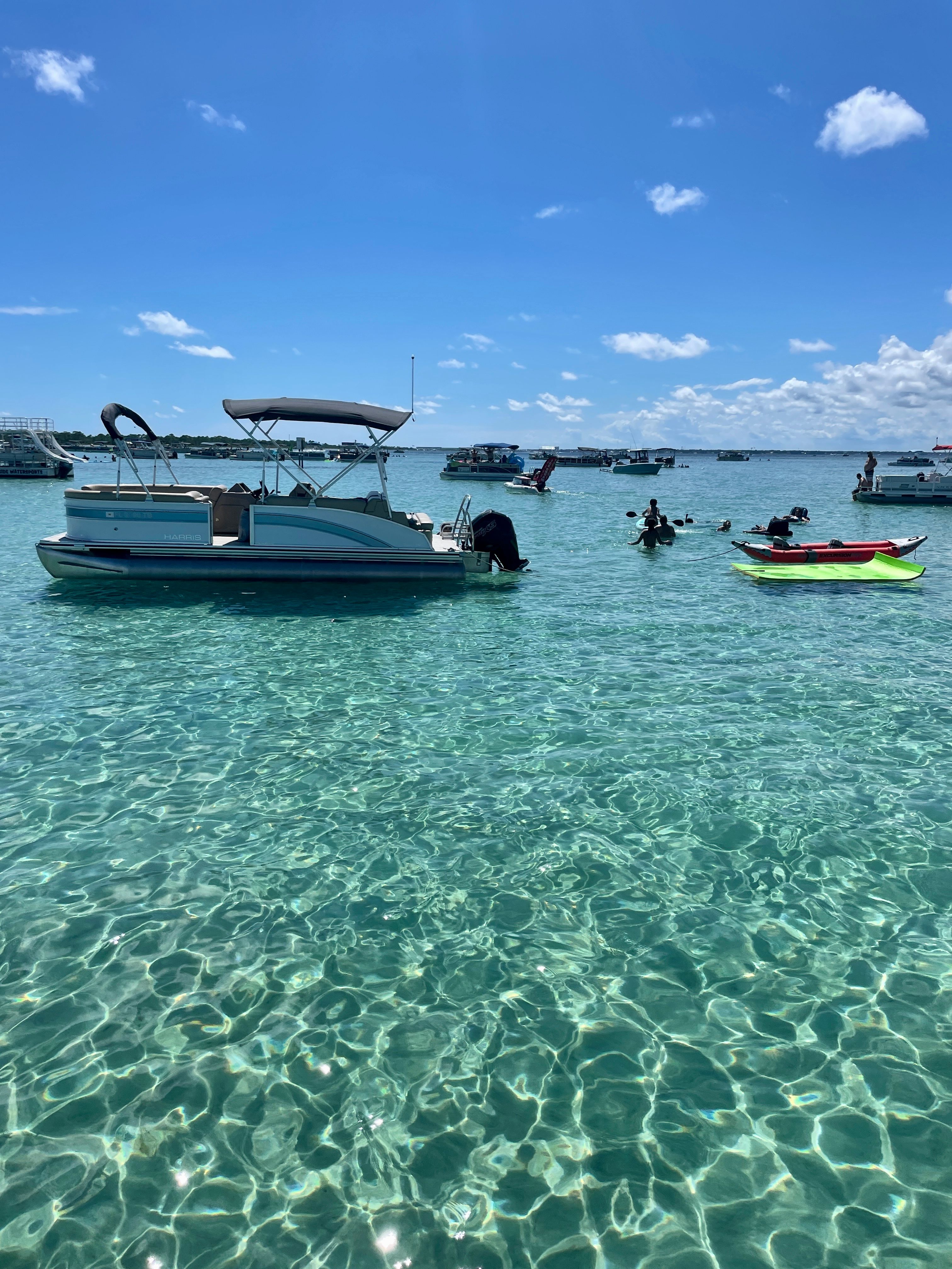 Pontoon boat and bright kayaks floating on sparkling turquoise water beneath a sunny blue sky, with other boats anchored in a shallow coastal bay