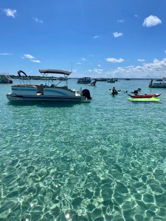 Pontoon boat and bright kayaks floating on sparkling turquoise water beneath a sunny blue sky, with other boats anchored in a shallow coastal bay