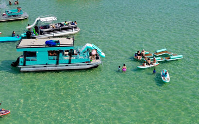 Aerial view of a turquoise pontoon houseboat with slide, surrounded by paddleboards, floating lounge platforms and people swimming in clear shallow turquoise coastal water.