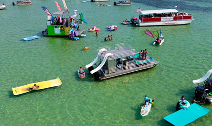 Aerial view of a sunny shallow turquoise bay with pontoon boats and a slide-equipped party barge, people on inflatable floats and paddleboards enjoying a lively summer boating day.
