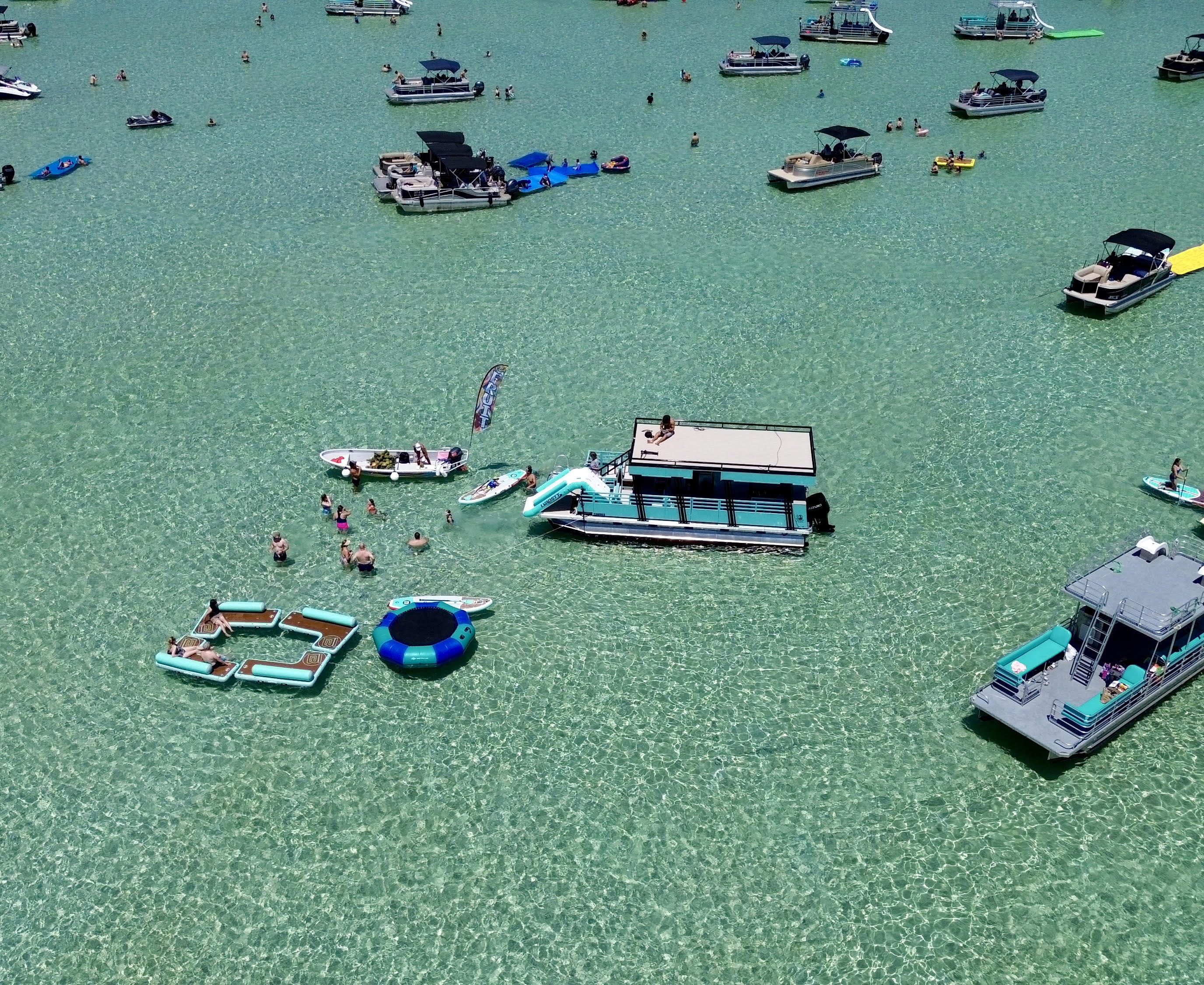 Aerial view of a sun-soaked shallow turquoise bay dotted with pontoon boats, paddleboards, inflatable loungers and swimmers enjoying a summer day.
