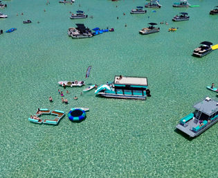 Aerial view of a sun-soaked shallow turquoise bay dotted with pontoon boats, paddleboards, inflatable loungers and swimmers enjoying a summer day.