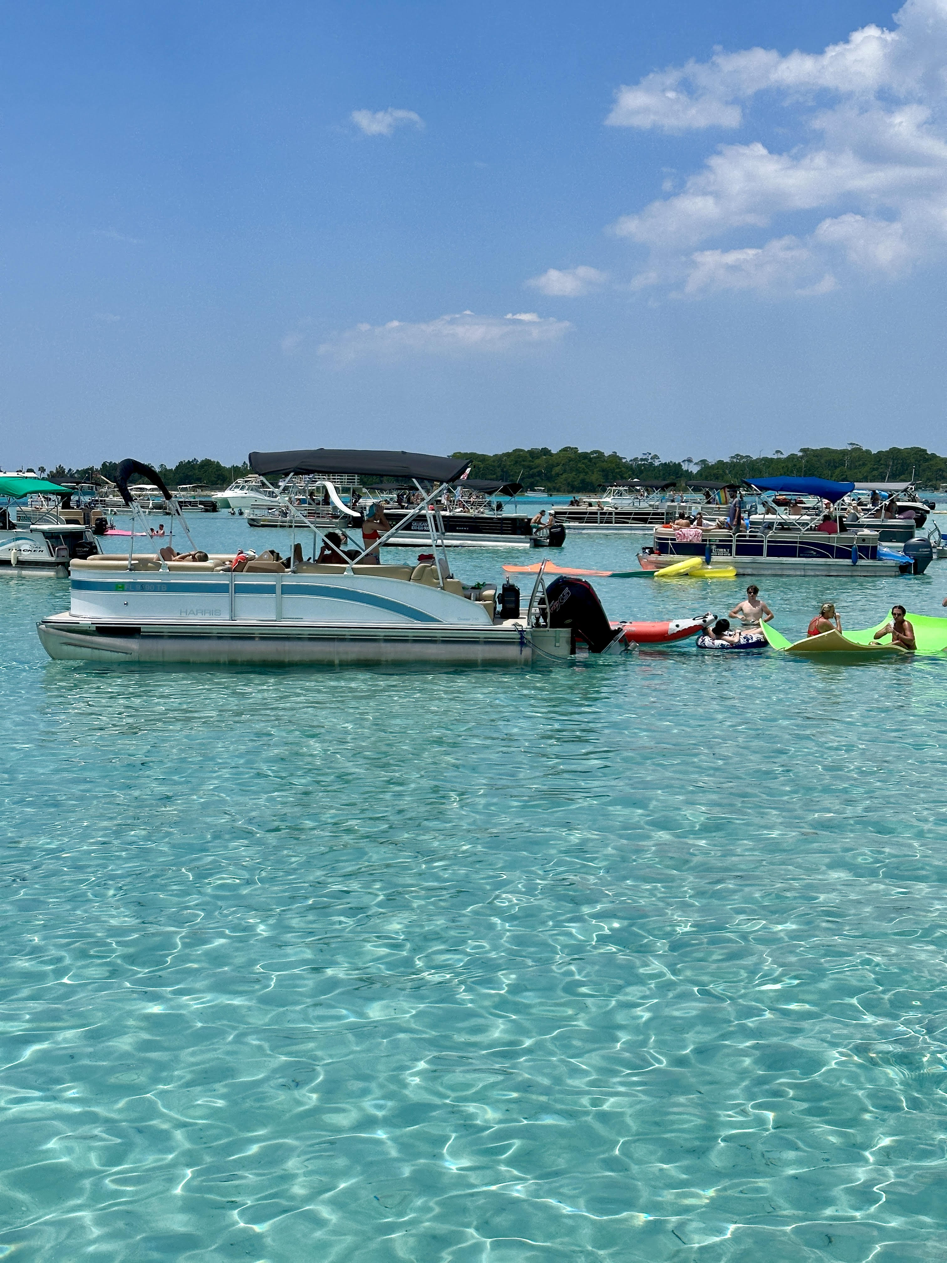 Sunny summer scene of pontoon boats and kayaks anchored in clear turquoise shallow water, people lounging on inflatables near a tree-lined shore under a bright blue sky.