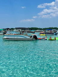 Sunny summer scene of pontoon boats and kayaks anchored in clear turquoise shallow water, people lounging on inflatables near a tree-lined shore under a bright blue sky.