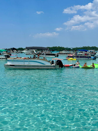 Sunny summer scene of pontoon boats and kayaks anchored in clear turquoise shallow water, people lounging on inflatables near a tree-lined shore under a bright blue sky.