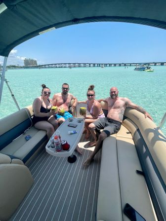 Four friends relaxing on a pontoon boat with pineapple drinks in clear turquoise shallow water on a sunny day, coastal bridge and beachfront buildings visible in the distance