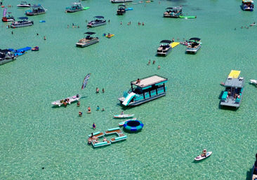 Aerial view of a sunny sandbar with clear turquoise water, dozens of pontoon boats, paddleboards and colorful floating inflatables with people swimming and relaxing.