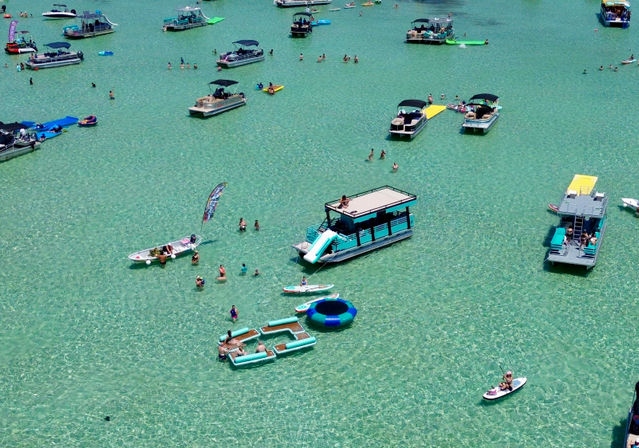 Aerial view of a sunny sandbar with clear turquoise water, dozens of pontoon boats, paddleboards and colorful floating inflatables with people swimming and relaxing.