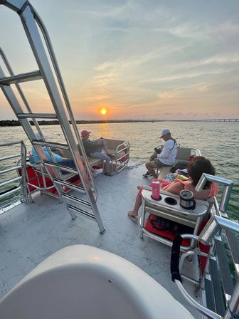 Group enjoying a relaxed sunset cruise on a pontoon boat, orange sun reflecting on calm coastal waters with a distant bridge and rocky jetty