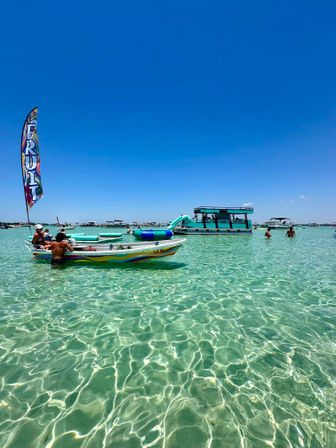 Sunny sandbar with crystal-clear turquoise water, a colorful small boat flying a tall "FRUIT" flag, swimmers nearby and a teal party pontoon with a waterslide in the background.