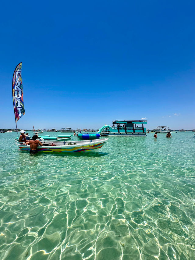 Sunny sandbar with crystal-clear turquoise water, a colorful small boat flying a tall "FRUIT" flag, swimmers nearby and a teal party pontoon with a waterslide in the background.