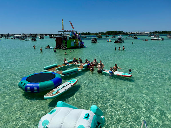 Sunny sandbar scene with people enjoying clear turquoise shallow water on inflatable trampolines, paddleboards and anchored boats under a bright blue sky.