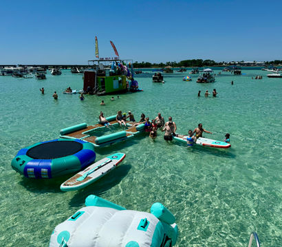 Sunny sandbar scene with people enjoying clear turquoise shallow water on inflatable trampolines, paddleboards and anchored boats under a bright blue sky.