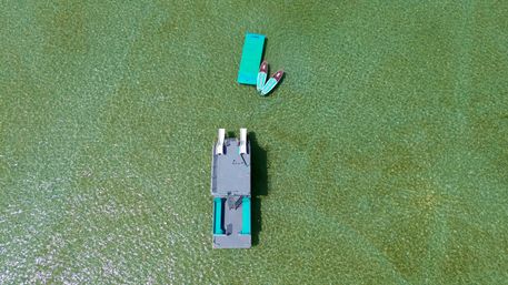 Aerial drone view of a teal-accented floating platform and two paddleboards drifting in shallow, crystal-clear green tropical lagoon water