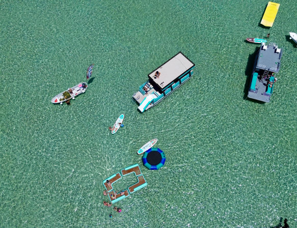 Aerial view of crystal-clear turquoise water with anchored party boats, paddleboards, a floating trampoline and inflatable lounge platforms, people relaxing and paddling.