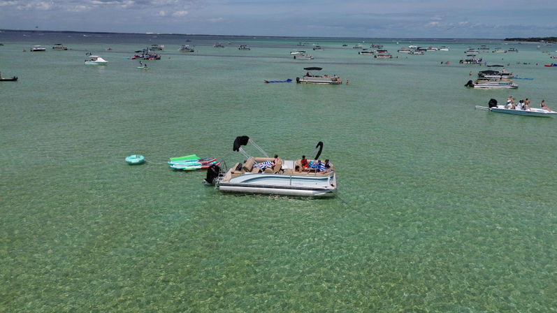 Pontoon boat anchored in shallow crystal-clear turquoise water with people lounging, inflatable floats and paddleboards nearby and dozens of boats scattered across the horizon under a sunny sky.