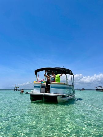 Pontoon boat anchored in shallow crystal-clear turquoise water with people aboard waving under a bright blue sky — sunny beach-day boating fun.