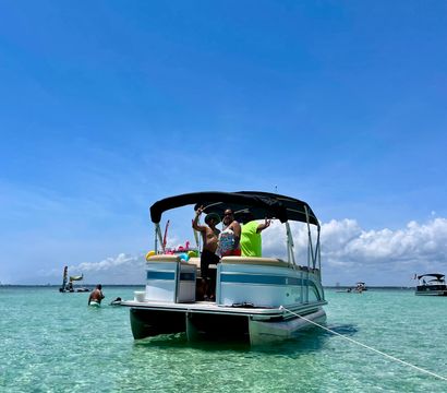 Pontoon boat anchored in shallow crystal-clear turquoise water with people aboard waving under a bright blue sky — sunny beach-day boating fun.