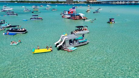 Aerial view of a sunny shallow turquoise bay with clear water, anchored party pontoons and boats with slides, paddleboards and floating mats, people swimming and relaxing near a bridge-lined shoreline.