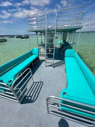 Pontoon boat interior with bright teal bench seating, stainless railings and ladder to an upper deck, anchored in clear shallow turquoise water under a sunny blue sky with other boats in the distance.