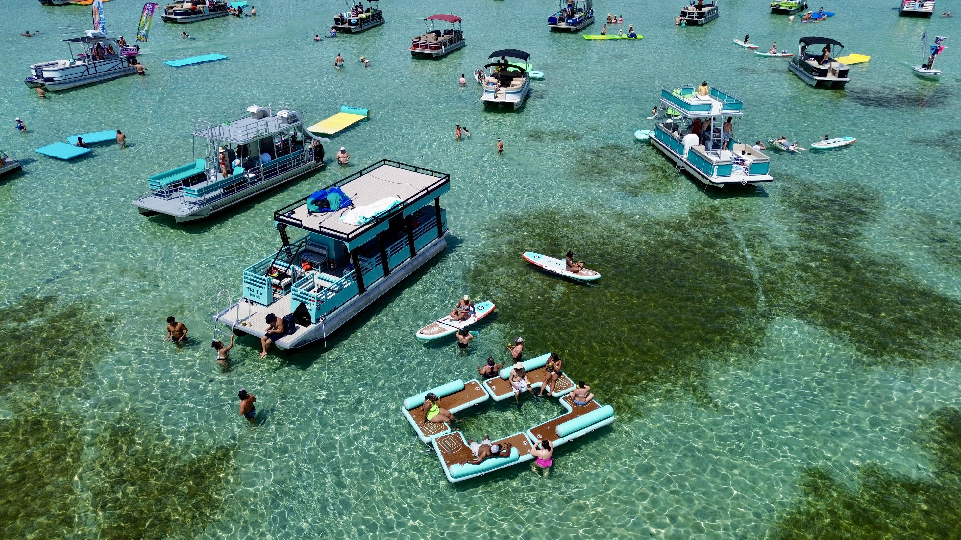 Aerial view of a lively sandbar party with pontoon boats, floating lounge platforms and paddleboards on crystal-clear turquoise water on a sunny summer day.